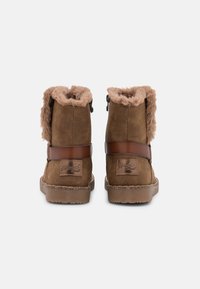 Botines de ante marrones con forro de pelo sintético, correa decorativa y parche de logo en la parte trasera. Suela de goma plana y cierre de cremallera lateral.