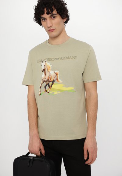 T-shirt à manches courtes vert olive avec un graphique d'un cheval au galop et le logo "Emporio Armani" imprimé au-dessus en beige. Modèle tenant un sac noir.
