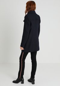 Kvinna med röd hår som bär en mörk marinblå kappa, svarta byxor med röda och vita sidstripe, samt svarta klackskor, stående mot en neutral bakgrund.