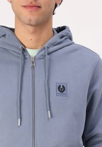 Modrá mikina na zips vyrobená z mäkkej tkaniny, s prednou vreckom, kapucňou so šnúrkou a čiernou nášivkou loga na ľavom prsníku. Strieborný zips ako detail.