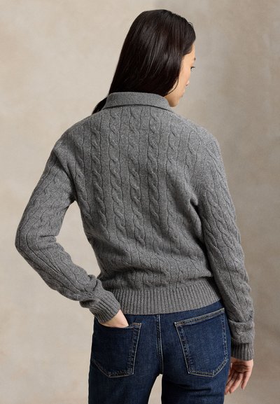 Femme aux longs cheveux foncés portant un pull en maille torsadée gris et un jean bleu, debout avec une main dans sa poche arrière.