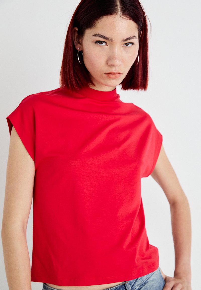 Czerwona bluzka z krótkim rękawem i wysokim kołnierzem, wykonana z gładkiego materiału. Posiada luźny krój i minimalne detalowanie szwów.
