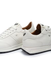 Zapatillas de piel blanca con un acabado suave, que presentan un logo negro en la lengüeta y una suela de goma texturizada. Diseño clásico con cordones.