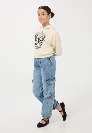Przycięta beżowa bluza z kapturem z czarnym graficznym motylem i napisem. Luźne, jasnoniebieskie dżinsy cargo z bocznymi kieszeniami. Czarne baleriny.