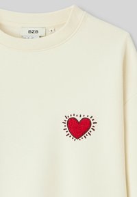 Sweatshirt couleur crème avec un patch en forme de cœur texturé rouge, bordé de noir et avec des lignes rayonnantes, situé sur la zone gauche de la poitrine.