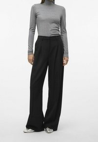 Top à col montant gris à manches longues associé à un pantalon noir à taille haute et jambes larges. Le modèle porte des chaussures plates argentées. Tissu lisse et ajusté.