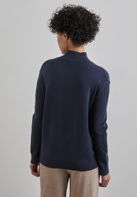 Marinblå stickad tröja med hög ribbad hals och ribbade ärmmuddar, med rak passform och mjuk textur. Bärs med beige kordflidbyxor.