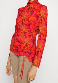 Röd långärmad topp med ett blommönster i rosa och grönt, med rynkning på sidorna och en hög krage. Matchas med beige texturerade shorts.