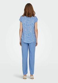 Donna in piedi di spalle, che indossa una maglietta a maniche corte blu con fiori, pantaloni azzurri chiari e scarpe con tacchi alti bianche, su uno sfondo neutro.