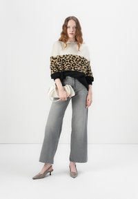 Leopardprint gebreide trui met een beige bovenhelft, zwarte manchetten; grijze wijd uitlopende jeans; lichtgrijze slingback hakken; houdt een kleine witte tas vast.