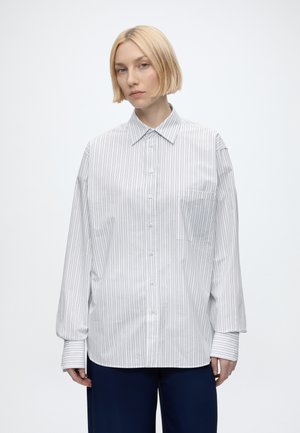 Blond persoon met een bobkapsel, draagt een losse witte blouse met dunne verticale strepen en donkere broek, staand tegen een egale achtergrond.
