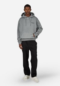 Pelēka kapucēta džemperis ar priekšējo kabatu un auklām, apvienots ar melnām biksēm. Balti sporta apavi pilnveido tērpu.