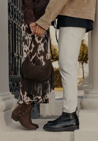 Hnědé semišové kotníkové boty s dekorativními popruhy, černé kožené šněrovací boty, hnědá kabelka s třásněmi a vzorovaná sukně s přírodními odstíny.