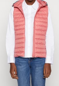 Padded ärmellose Weste in Koralle, mit durchgehendem Reißverschluss, feinen horizontalen Steppnähten und einer Kapuze mit blauen Akzenten. Getragen über einem weißen Shirt.