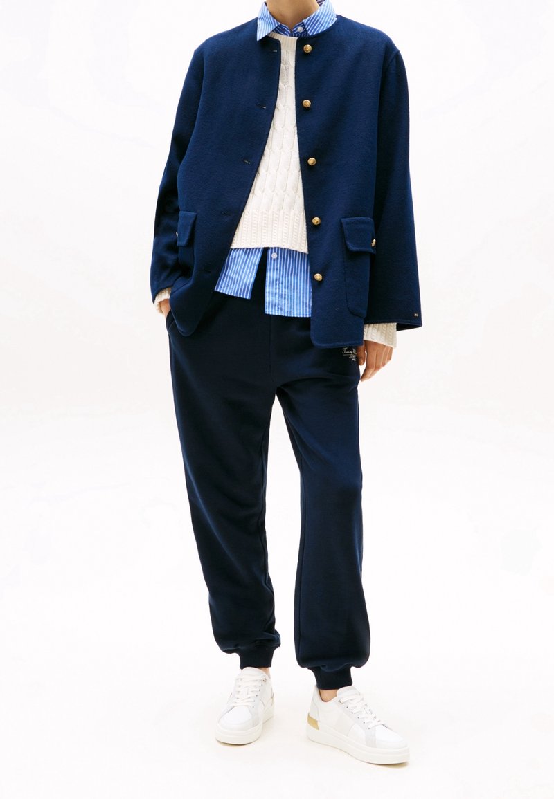 Personne portant une veste bleu marine à boutons dorés, un pull blanc en maille torsadée, une chemise rayée bleue, un jogging bleu marine et des baskets blanches.