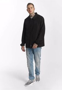 Czarna kurtka z guzikami, jasnoszara koszula pod spodem, jasnoniebieskie przetarte dżinsy, szare obuwie sportowe. Kurtka ma logo.