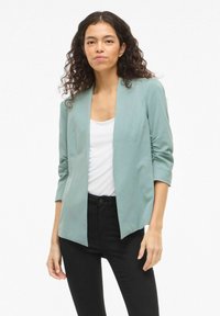 Femme aux cheveux bouclés portant un blazer vert clair à ouverture devant, un haut blanc et un pantalon noir, se tenant devant un fond blanc.