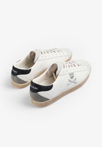 Zapatillas de cuero blanco con contrafuerte texturizado en gris y negro, que presentan un detalle de calavera y tibias cruzadas. Suela de goma color beige claro.