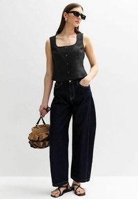 Top court noir à encolure carrée avec détails en boutons, associé à un jean large bleu foncé. Le modèle tient un sac en osier.