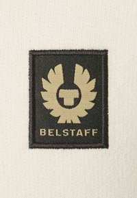 Svart tyg med guldbroderi som visar en bevingad emblem och texten "BELSTAFF" längst ner, mot en krämfärgad bakgrund.