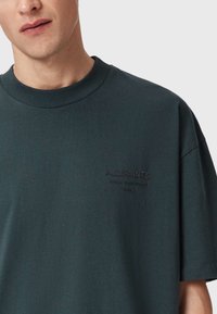 Ciemnozielona bawełniana koszulka z okrągłym dekoltem, w luźnym fasonie, z tłoczonym logo "AllSaints" na piersi. Gładka faktura i krótkie rękawy.