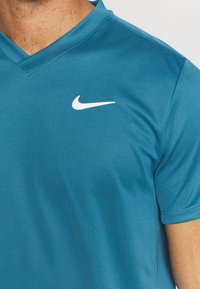 Petrolejová sportovní polo košile ze strukturované tkaniny, má design v výstřihu do V a bílé logo Nike v levém horním rohu.