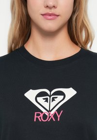 Fekete póló, fehér szív és gyémánt logóval, alatta rózsaszín "ROXY" felirattal. Kerek nyakkivágás és puha pamut anyag.