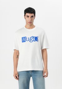 Camiseta de manga corta blanca con un diseño gráfico en texto azul "HugO" en varios estilos. Tejido suave con un ajuste relajado.