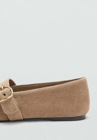 Jasnobrązowy, płaski but z zamszu z złotą klamrą i czarną podeszwą, pokazany z boku na jasnym, jednolitym tle.