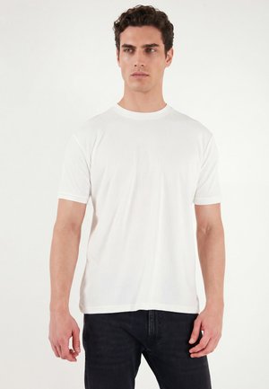 Mand iført en simpel hvid kortærmet T-shirt og sorte bukser, stående foran en ensfarvet hvid baggrund, kigger let til siden.