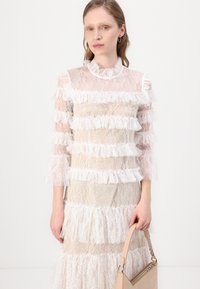 Rochie alba din dantelă cu volane stratificate și mâneci trei sferturi, având un căptușel moale bej și un gât înalt. Accentuata cu o geantă nude.