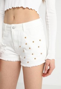 Bílé džínové šortky s třepícím se lemem, zdobené zlatými cvočky na přední straně a mírně opraným vzhledem na jedné straně.