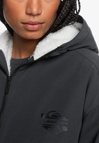 Čierna kapucňová bunda s bielou, nadýchanou podšívkou. Má zips a čierne logo na ľavej hrudi. Mäkšia textúra.