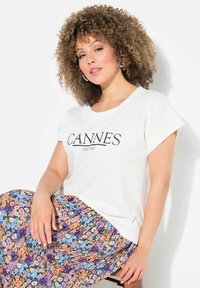 Camiseta de manga corta Beige claro con "CANNES DAY TRIP" impreso en negro. Una falda plisada de flores multicolores presenta tonos de púrpura, azul y naranja.