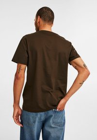 Brun bomulds t-shirt med korte ærmer, rund hals og afslappet pasform. Båret med lyseblå denimbukser, der viser en tatovering på armen.