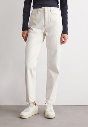 Person wearing white high-waist jeans and white sneakers, standing on a gray floor against a plain white wall.