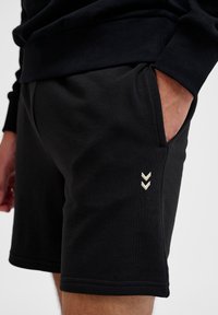 Mustat shortsit, jotka on valmistettu pehmeästä kankaasta. Niissä on sivutasku ja koristeellinen vaaleankermanvärinen viivasto-logo vasemmalla puolella.