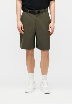 Mees, kes kannab oliivrohelisi põlvpikkusi lühikesi pükse musta võrguga vööga, valgeid sokke ja valgeid tossusid, seisab ühevärvilise tausta ees.