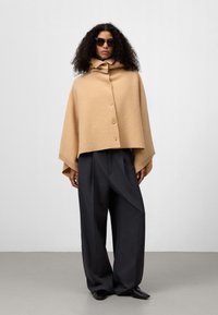 Cappotto in lana beige con colletto alto, chiusura a bottone e maniche ampie, indossato sopra pantaloni grigi a gamba larga e scarpe nere.