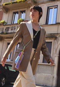 Blazer marrón con solapas blancas, top negro, pantalones beige y un bolso bandolera colorido con una mezcla de azul claro, púrpura y texturas de madera.