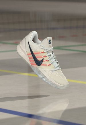 Chaussure de sport blanche avec un logo Nike noir et des rayures orange, lacets noués, suspendue au-dessus du sol d'un terrain de sport intérieur.