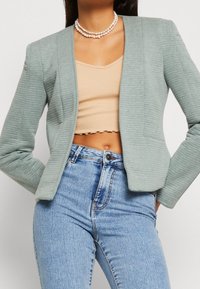 Blazer bleu clair côtelé avec un design structuré, col cranté et silhouette ajustée. Porté sur un crop top beige et associé à un jean taille haute.
