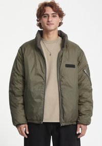 Chaqueta acolchada verde oliva con cierre frontal, puños elásticos y cinturilla. Presenta un parche de logo en el lado izquierdo del pecho y un bolsillo con cremallera.