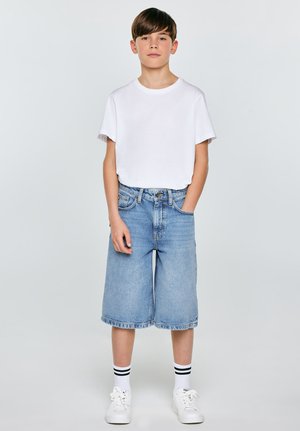 Chlapec stojící, oblečený v bílém tričku, dlouhých džínových šortkách, bílých teniskách a bílých ponožkách s dvěma černými pruhy, ruce uvolněné, neutrální pozadí.