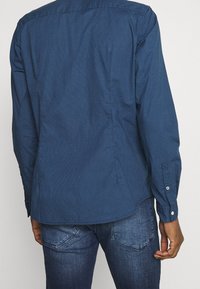 Camicia a maniche lunghe blu scuro con un piccolo motivo geometrico, caratterizzata da un colletto con bottoni e una vestibilità sartoriale, abbinata a jeans di denim.