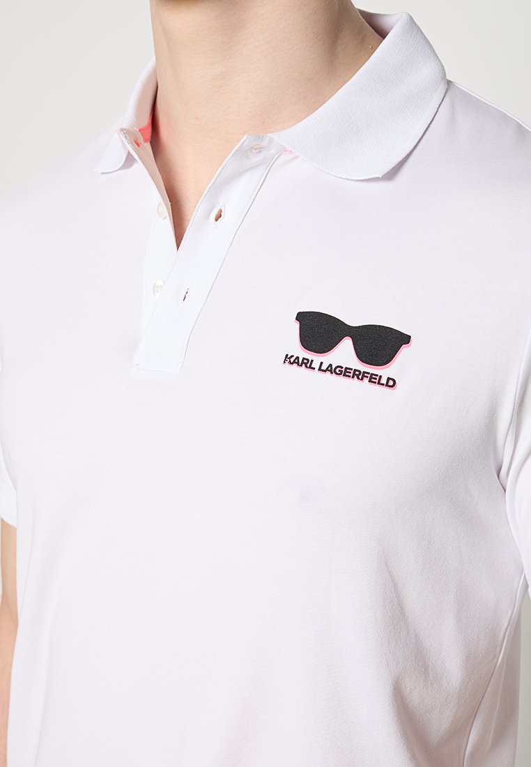Biała koszulka polo z kołnierzykiem i guzikami, z czarnym logo okularów przeciwsłonecznych oraz napisem "KARL LAGERFELD" na klatce piersiowej.