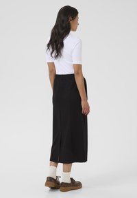 Jupe midi noire avec fente arrière, taille élastique et texture lisse. Associée à un haut blanc à manches courtes et des chaussures marron avec des chaussettes crème.