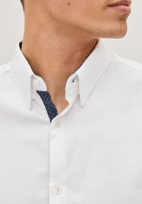 Camisa blanca de botones con un cuello estampado en azul oscuro en contraste. Presenta una textura suave y un diseño de cuello estándar con dos botones.