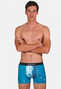 Shorts de bain bleus présentant un motif de vagues, une taille élastique avec le logo "WAXX" et un design ajusté qui met en valeur la silhouette du corps.