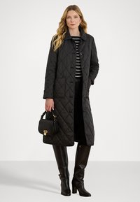 Zwarte gewatteerde lange jas met knoopsluiting, twee voorkruiken en een klassieke kraag. Gecombineerd met een gestreepte top, zwarte broek en kniehoge laarzen.